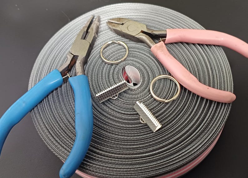 May include: Two pairs of pliers, one blue and one pink, are lying on a roll of black and white striped ribbon. Two silver metal clasps are also on the ribbon.