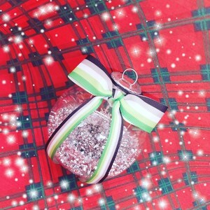 Op de afbeelding: Een transparante glazen kerstbal gevuld met zilveren glitter. Hij is versierd met een groen, wit en zwart lint dat in een strik is gebonden.