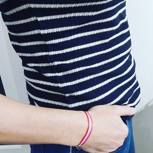 Pode incluir: Uma pessoa vestindo uma camisa listrada preta e branca e jeans azul com uma pulseira listrada rosa, branca e vermelha no pulso.