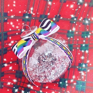 Op de afbeelding: Een transparante glazen kerstbal gevuld met zilveren glitter. De bal is versierd met een regenboogkleurig en zwart-wit gestreept lint.