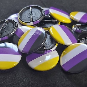 Peut inclure: Gros plan sur un tas de boutons ronds à épingle avec un motif rayé jaune, blanc et violet. Les boutons sont disposés de manière aléatoire.