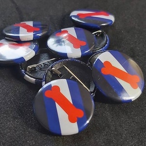 May include: A set of seven blue, white and red pinback buttons with a red bone graphic. The buttons are arranged in a pile on a black surface.