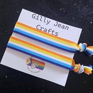 May include: Two elastic hair ties featuring horizontal stripes of orange, yellow, white, and blue. The hair ties are attached to a white display card printed with the text Gilly Jean Crafts and a logo featuring a unicorn head and rainbow colors.