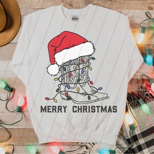 Puede incluir: Sudadera blanca con un dibujo de una bota vaquera con un gorro de Pap&aacute; Noel y envuelta en luces de Navidad. El texto "Merry Christmas" est&aacute; impreso debajo de la bota.