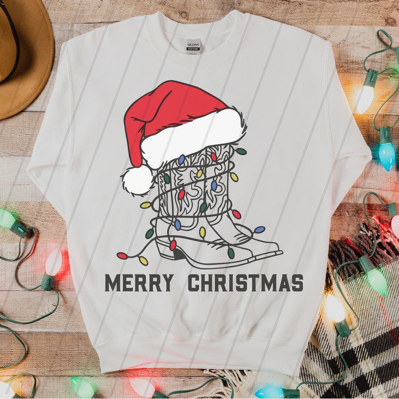 Puede incluir: Sudadera blanca con un dibujo de una bota vaquera con un gorro de Pap&aacute; Noel y envuelta en luces de Navidad. El texto "Merry Christmas" est&aacute; impreso debajo de la bota.