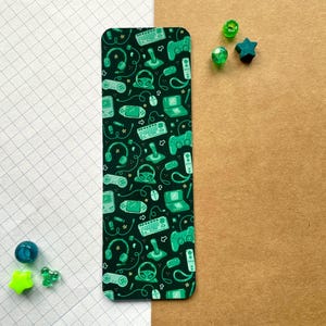 May include: A green and black bookmark featuring a repeating pattern of video game controllers, game cartridges, and other gaming accessories.