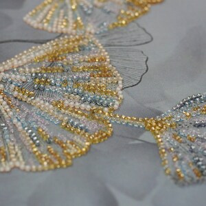 May include: Close-up of a beaded fabric with a design of ginkgo leaves. The leaves are made of small, iridescent beads in shades of gold, silver, and blue.