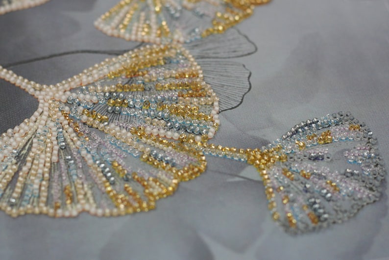 May include: Close-up of a beaded fabric with a design of ginkgo leaves. The leaves are made of small, iridescent beads in shades of gold, silver, and blue.