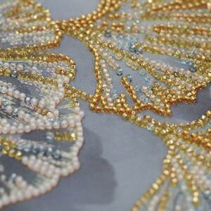 May include: Close-up of a fabric with intricate beading in shades of gold, white, and blue. The beads are arranged in a pattern that resembles a floral design.