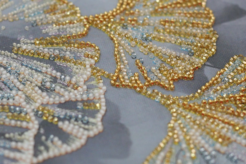 May include: Close-up of a fabric with intricate beading in shades of gold, white, and blue. The beads are arranged in a pattern that resembles a floral design.