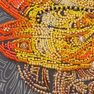 May include: Close-up of a beaded artwork depicting a stylized sun with orange, yellow, and gold beads. The beads are arranged in a swirling pattern, creating a sense of movement and energy.