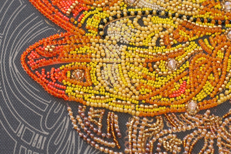 May include: Close-up of a beaded artwork depicting a stylized sun with orange, yellow, and gold beads. The beads are arranged in a swirling pattern, creating a sense of movement and energy.