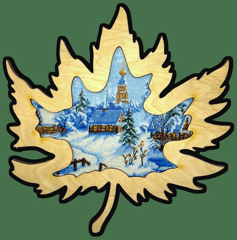 May include: A wooden maple leaf cutout with a winter village scene inside. The scene features a church, houses, and snow-covered trees.