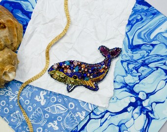 Op de afbeelding: Een blauwe en paarse walvis broche met gouden en bruine accenten. De walvis is gemaakt van kralen en pailletten en is een grillig en kleurrijk sieraad.