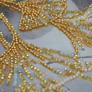 May include: Close-up of a fabric with gold and white beads sewn in a pattern. The beads are arranged in a floral design.