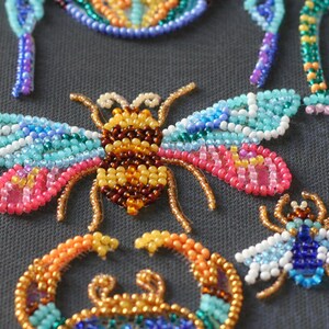 May include: A close-up of a beaded bee with brown, yellow, and orange beads for the body and blue, pink, and white beads for the wings. The bee is surrounded by other beaded insects, including a dragonfly and a crab.