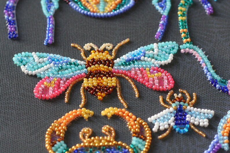 May include: A close-up of a beaded bee with brown, yellow, and orange beads for the body and blue, pink, and white beads for the wings. The bee is surrounded by other beaded insects, including a dragonfly and a crab.
