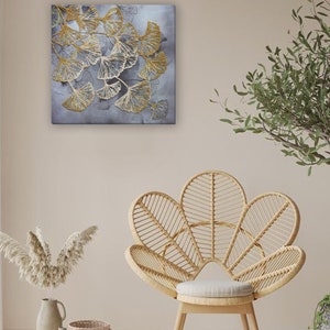 May include: A square canvas wall art featuring a stylized design of gold ginkgo leaves on a gray background. The art is displayed on a wall with a wicker chair and a vase of dried pampas grass.