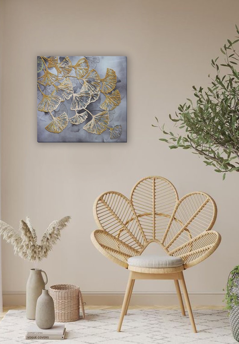 May include: A square canvas wall art featuring a stylized design of gold ginkgo leaves on a gray background. The art is displayed on a wall with a wicker chair and a vase of dried pampas grass.