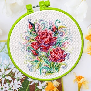 Può includere: Un cerchio da ricamo verde con una tela bianca che presenta un disegno a punto croce di rose rosse e rosa con foglie e steli verdi.