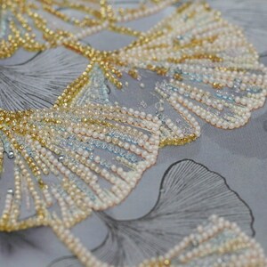 May include: Close-up of a fabric with a beaded design of ginkgo leaves. The leaves are made of gold, white, and blue beads. The fabric is a light gray color.