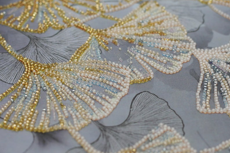 May include: Close-up of a fabric with a beaded design of ginkgo leaves. The leaves are made of gold, white, and blue beads. The fabric is a light gray color.