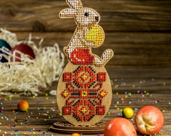 Può includere: Una decorazione per uova di Pasqua in legno con un motivo geometrico colorato. Un coniglio marrone che tiene un uovo giallo è seduto sopra l'uovo. La decorazione è circondata da spolverate colorate.