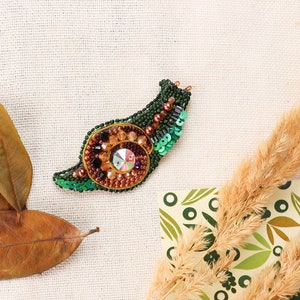 May include: A green and brown beaded snail brooch with a sparkly iridescent center. The brooch is on a beige fabric background.