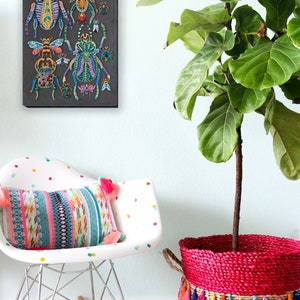 May include: A colorful beaded art print of various insects, including beetles and butterflies, on a black background. The print is hanging on a light blue wall above a white rocking chair with a colorful patterned pillow. A potted plant with green leaves is in a wicker basket with a pink base and colorful tassels.