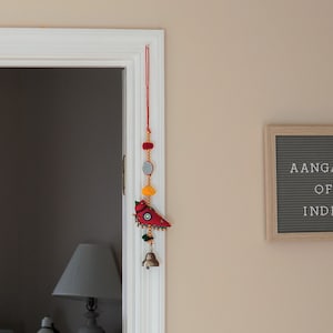 May include: A white door frame with a red and yellow hanging decoration with a bird design. The door opens to a room with a window and two laptops on a desk. A sign on the wall reads "Aangan of India".