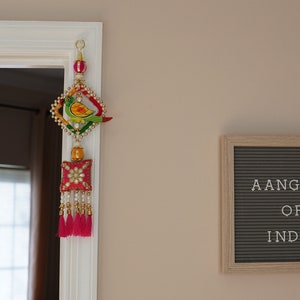 May include: A colorful door hanging with a bird design and pink tassels. The hanging is decorated with beads and sequins. A letter board with the text "Aangan of India" is on the wall.
