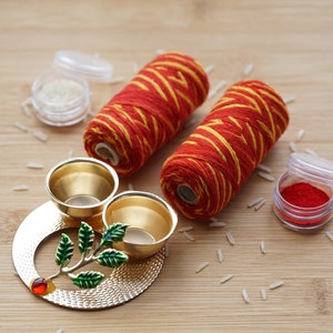 May include: A gold metal tray with two small bowls and a decorative leaf design. The tray is surrounded by red and yellow thread spools, small containers of white and red powder, and scattered white rice.