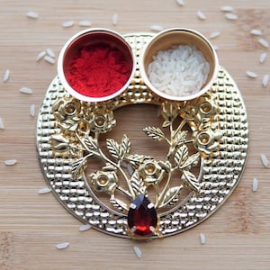 May include: A gold metal tray with a floral design and two small bowls. One bowl contains red powder and the other contains white rice. A red gemstone is in the center of the tray.