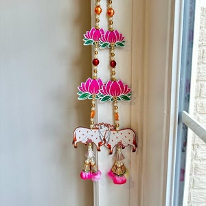 May include: A pink and green lotus flower garland with white beads and gold accents. The garland features two white cows with pink and gold details. The garland is hanging from a white door frame.
