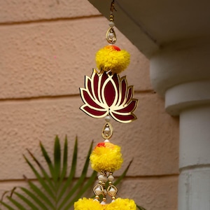 May include: A decorative hanging ornament featuring a lotus flower design in gold and burgundy, accented with yellow pom-poms and pearl-like beads. The ornament is suspended from a hook, creating a festive and elegant display.