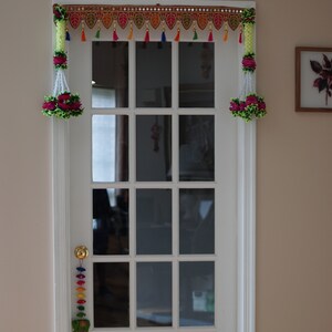 May include: A white door with a glass pane and a colorful garland hanging above it. The garland has green, red, yellow, and blue tassels. The door has a gold door handle.