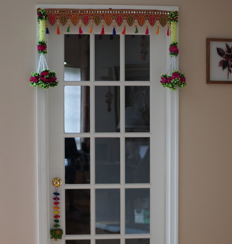 May include: A white door with a glass pane and a colorful garland hanging above it. The garland has green, red, yellow, and blue tassels. The door has a gold door handle.