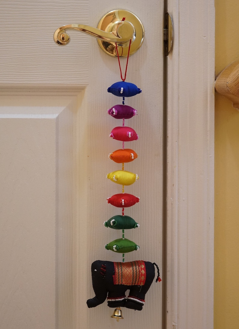 May include: A rainbow-colored fabric garland with a black elephant charm hanging from a door knob. The garland is made of small, round fabric pieces sewn together with white beads.