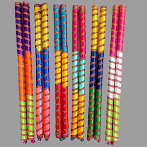 May include: A collection of colorful, decorative wooden sticks. Each stick is wrapped in vibrant ribbons of orange, purple, yellow, blue, green, and pink. The sticks are adorned with gold-colored accents and are arranged vertically against a neutral background.