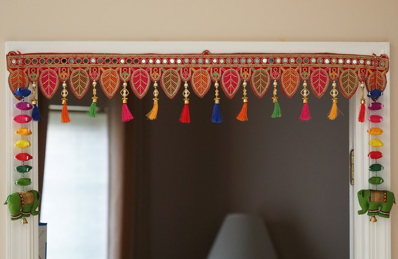 May include: A colorful door hanging with red fabric, gold trim, and mirrored accents. It features a row of tassels in various colors, including blue, green, orange, and yellow. The hanging is decorated with small, beaded leaves.