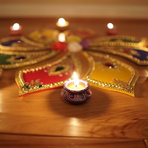 May include: A colorful rangoli design with lit tealight candles in small, decorative bowls. The design features intricate patterns and bright colors, including red, yellow, green, and blue.