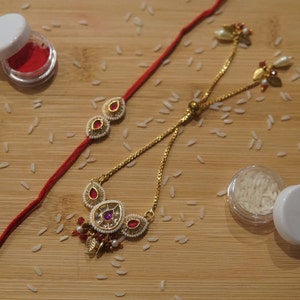 May include: A gold-toned necklace and bracelet set with red and white accents. The jewelry features a teardrop design with red gemstones and white pearls. The set is displayed on a wooden surface with scattered rice grains and small containers.