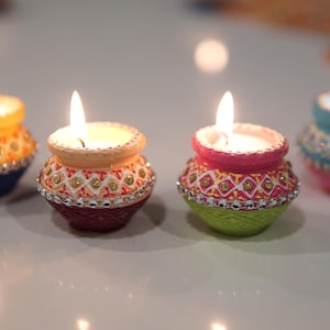 May include: Four small, decorative candles in colorful, ornate pots. The pots are painted in shades of yellow, blue, pink, and green, with intricate patterns and sparkling embellishments. Each candle is lit, casting a warm glow.