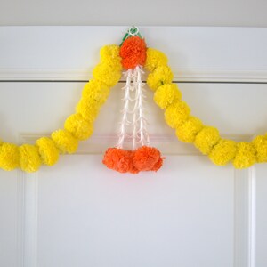 K&ouml;nnte beinhalten: Eine gelbe Ringelblumenkette mit orangefarbenen und wei&szlig;en Bl&uuml;tenakzenten h&auml;ngt an einer wei&szlig;en T&uuml;r.
