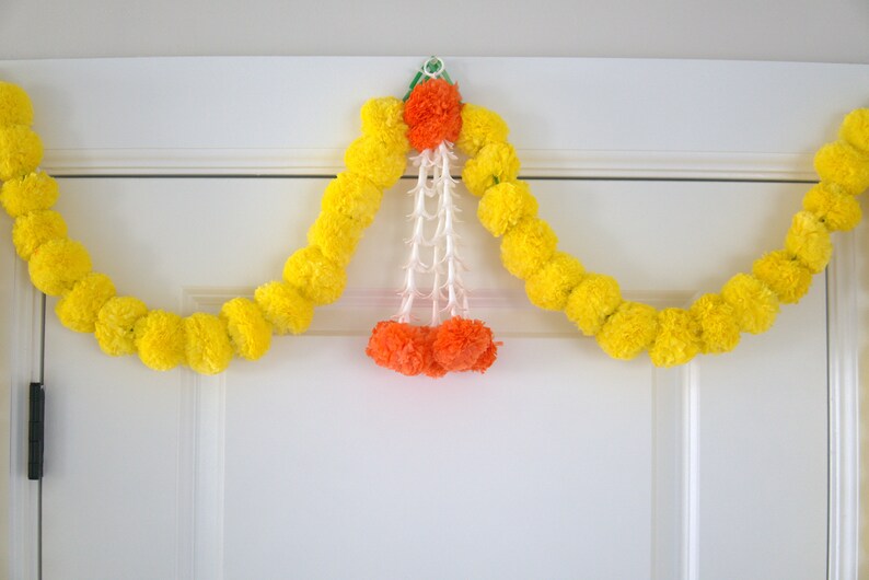 K&ouml;nnte beinhalten: Eine gelbe Ringelblumenkette mit orangefarbenen und wei&szlig;en Bl&uuml;tenakzenten h&auml;ngt an einer wei&szlig;en T&uuml;r.