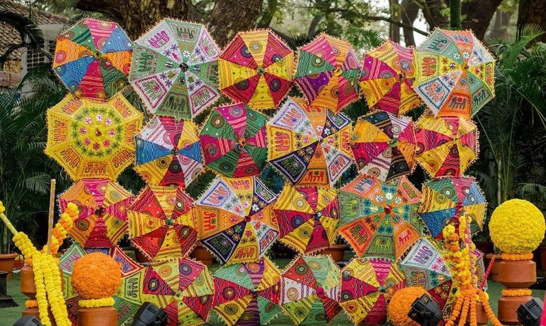 May include: A vibrant display of colorful, patterned umbrellas. The umbrellas feature intricate designs in shades of yellow, orange, green, and blue. The scene is decorated with marigold garlands and spherical floral arrangements.