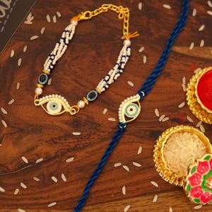 May include: A gold-toned bracelet featuring braided white and blue beads with evil eye charms and a gold-toned chain, alongside a blue braided thread bracelet with a central evil eye charm, both accented with small white pearls and gold hardware.