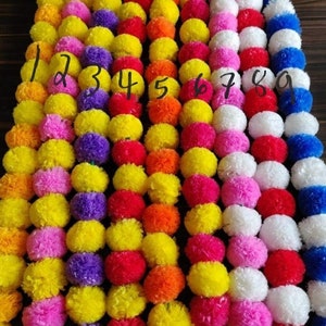 May include: Nine strands of colorful pom pom garland, each strand is a different color: orange, yellow, pink, purple, red, orange, yellow, white, and blue. The strands are numbered 1 through 9.