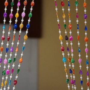 May include: A colorful beaded curtain made of iridescent teardrop-shaped beads strung on gold thread. The beads are arranged in a repeating pattern of different colors, including pink, blue, green, orange, and red.