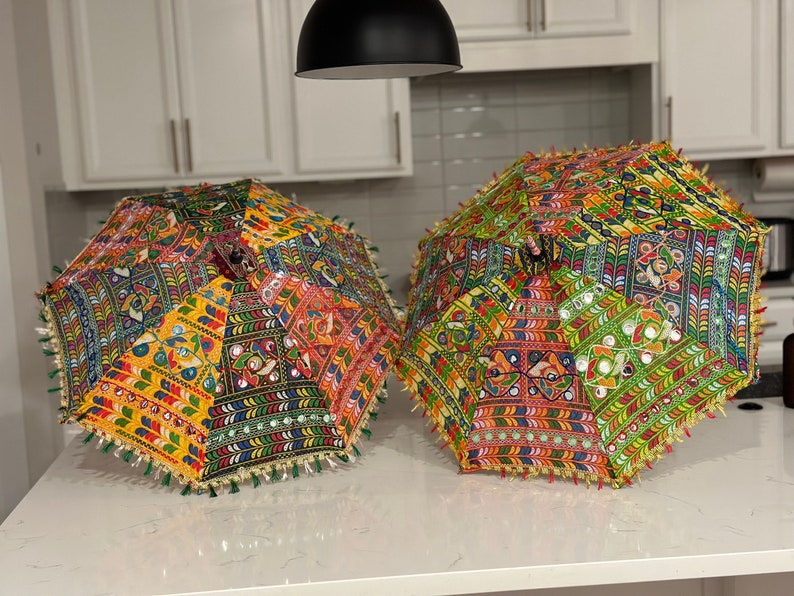 May include: Two colorful, decorative umbrellas with intricate patchwork designs. The umbrellas feature a vibrant mix of colors, including yellow, red, green, and blue, with mirrored accents and fringe trim. These umbrellas are perfect for adding a touch of bohemian flair.